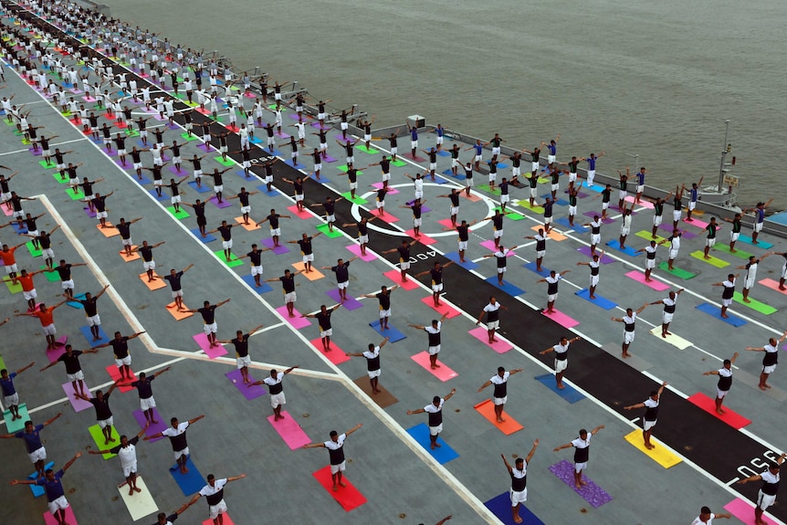 Thousands of people stand doing a yoga pose on the deck of an aircraft carrier.