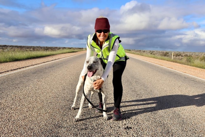 A woman wearing a hi vis vest poses for a photo in the middle of country road with a spotty black and white dog before her