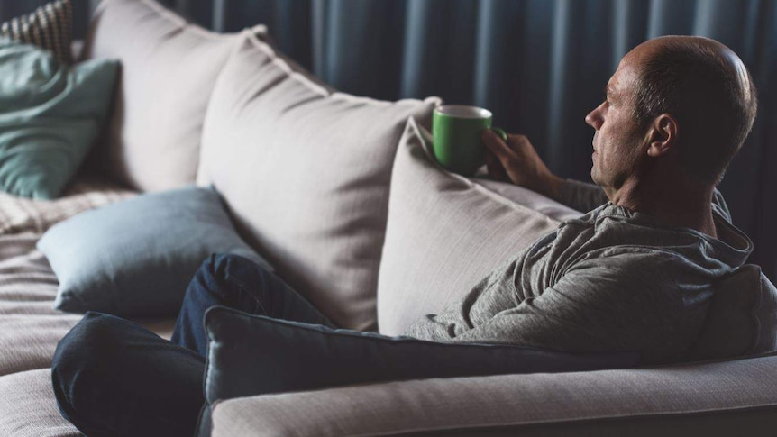 In a darkened room, a man sits cross-legged on a couch holding a green mug, looking into the distance.