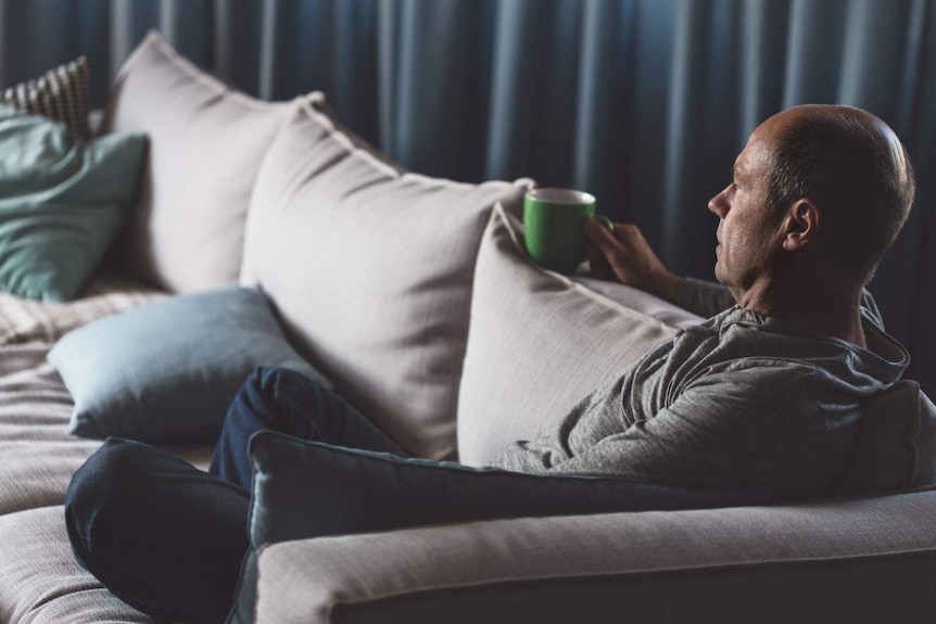 In a darkened room, a man sits cross-legged on a couch holding a green mug, looking into the distance.