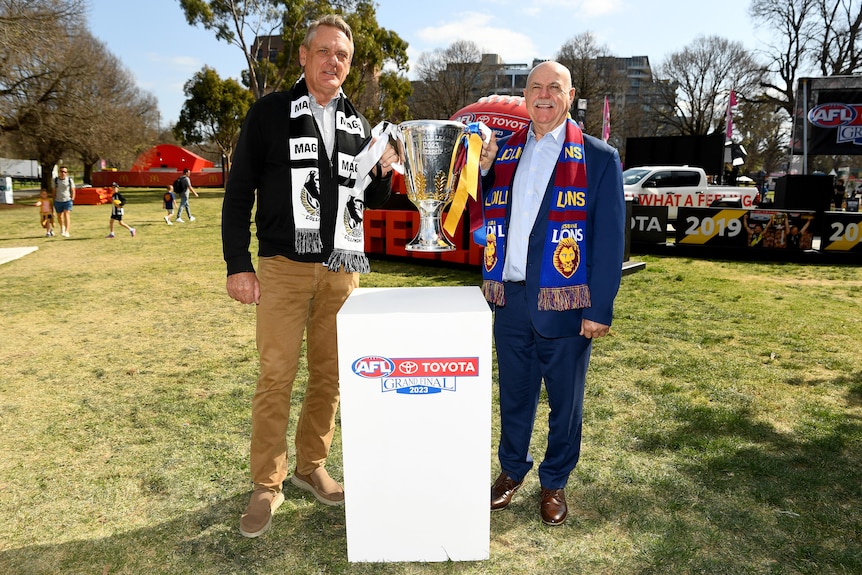 Two men, one wearing a Magpies scarf and the other with a Lions scarf stand holding a large silver cup over a pedestal.