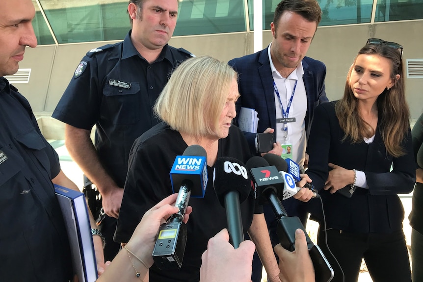 A frail woman stands over her walker in front of media.