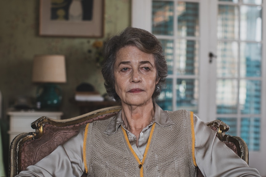 A glum-looking woman with gray hair wearing a gray silk shirt sits in an ornate chair