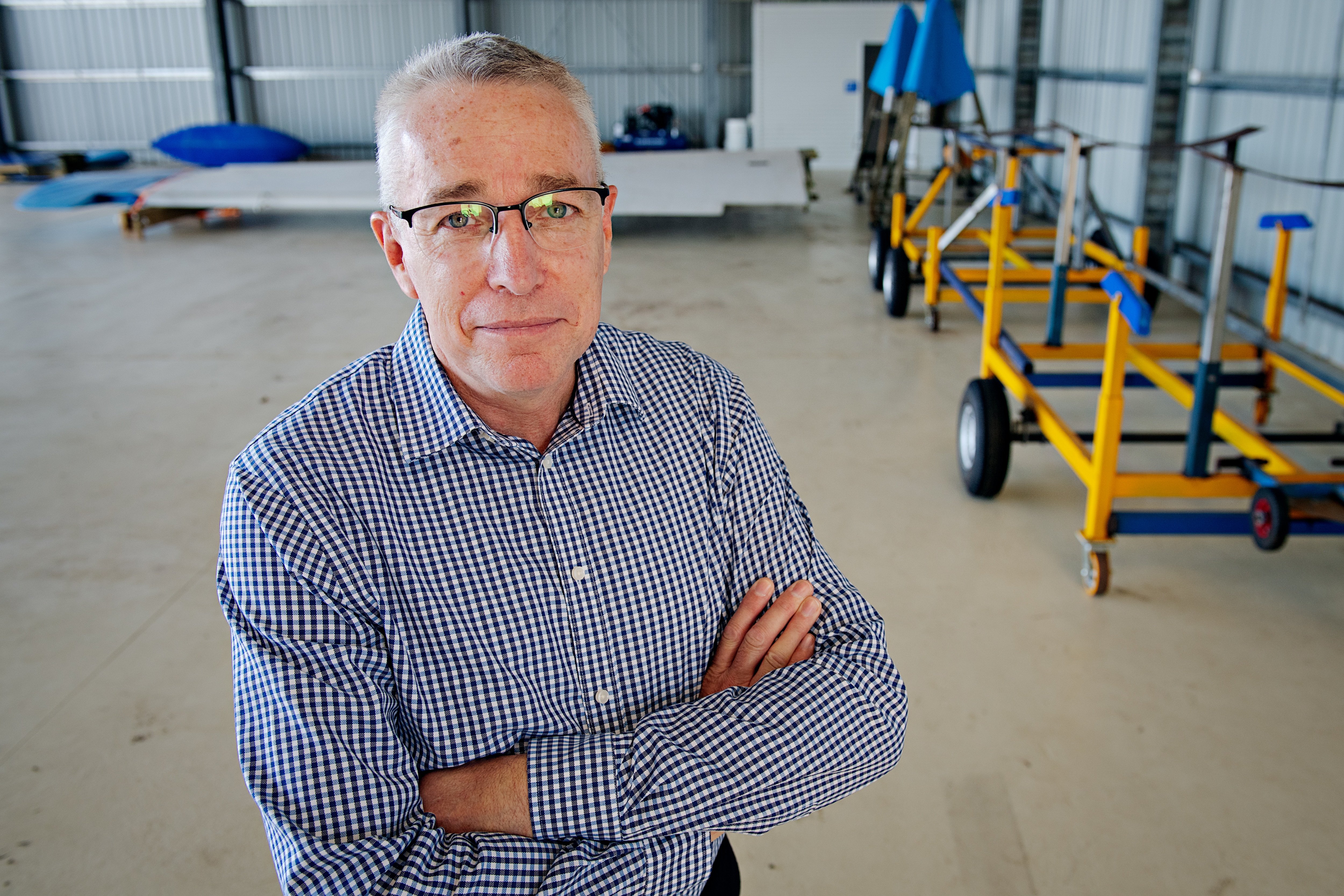 Un hombre blanco con una camisa a cuadros azules y blancas, pelo gris corto, gafas y un cobertizo al fondo.