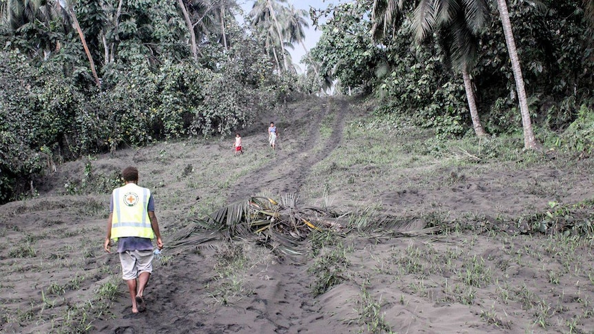 A red cross worker walks up an ash-covered hill as two villagers walk down.