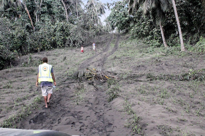 A red cross worker walks up an ash-covered hill as two villagers walk down.