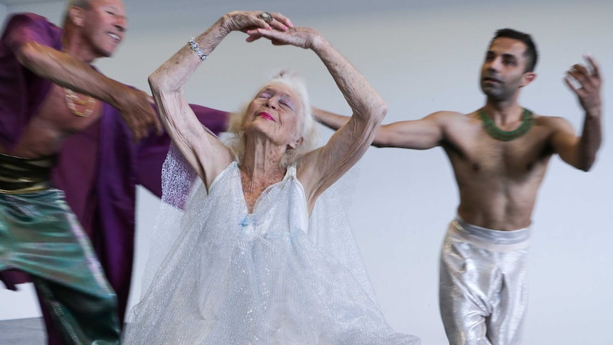 103-year-old Eileen Kramer in a glittering silver dress dances while sitting with two male dancers in the background