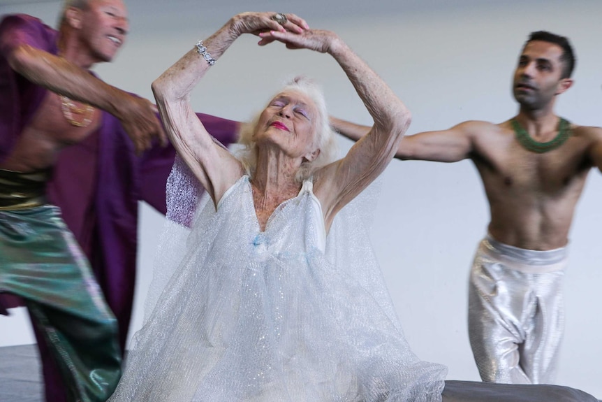 103-year-old Eileen Kramer in a glittering silver dress dances while sitting with two male dancers in the background