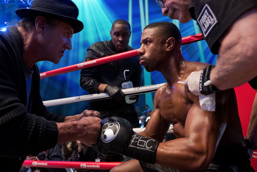 Colour still of Sylvester Stallone talking to Michael B. Jordan mid-match in 2018 film Creed II.