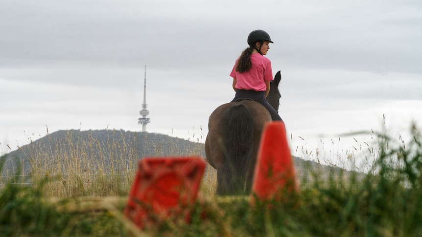 A young girl rides a brown horse with Telstra Tower in the background.