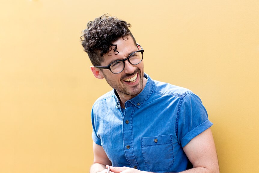 Caleb Rixon laughs, dressed in a blue shirt, in front of a yellow wall.