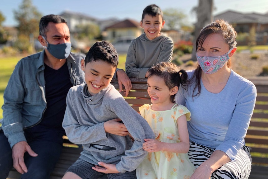 A young girl tickles her brother, as a family of five pose on a park bench.