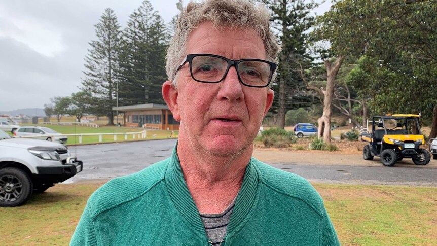 An older, bespectacled man in a green zip-up jumper stands at a beachside carpark.