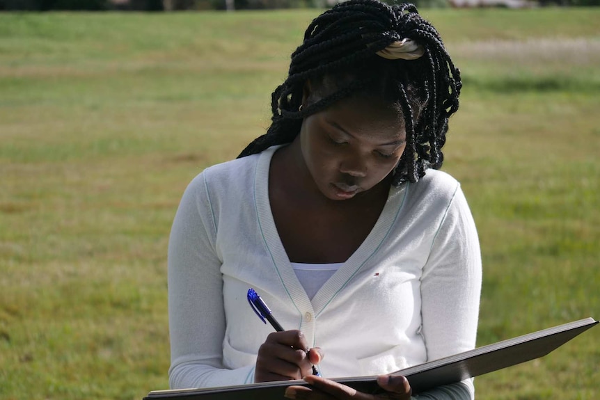A young woman sits on the grass, writing in a large book.