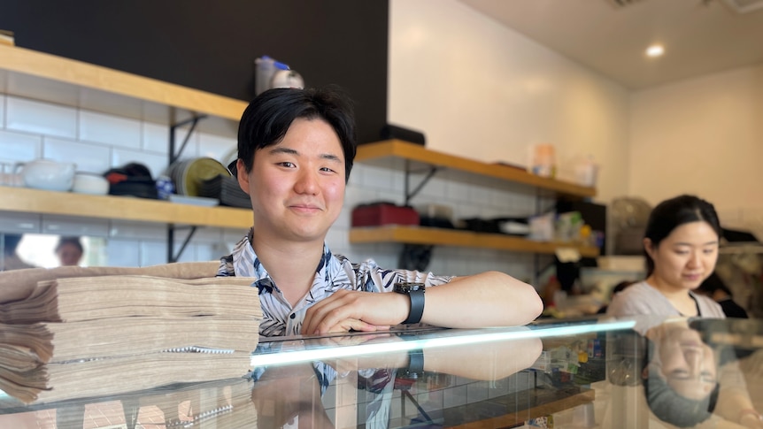 A young man with short black hair smiling behind a shop counter.