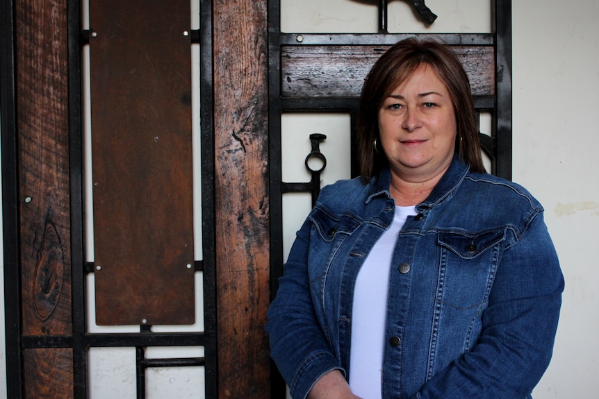 A woman in a white shirt and denim jacket stands in front of a wall with a wooden planks and metal on it.