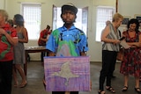 A boy standing in the middle of an art gallery holds up his painting of an eagle with a barramundi in its talons.