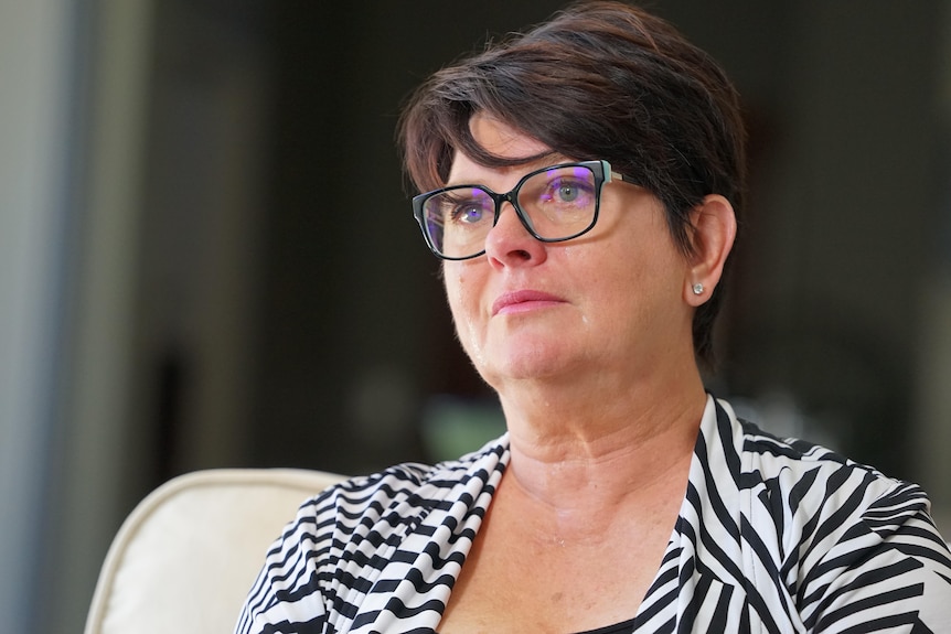 A middle-aged woman with short hair and glasses is being interviewed for TV. She wears a serious expression.