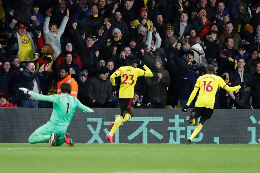 Footballers run in celebration toward the crowd after a goal, while the goalkeeper is left dejected.