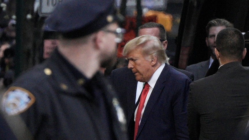 Donald Trump walks through a crowd of people, his head down. A police officer stands in the foreground.
