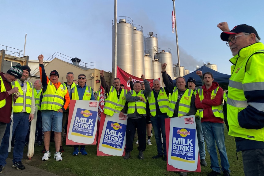 Men in high vis with signs stand outside a factory.