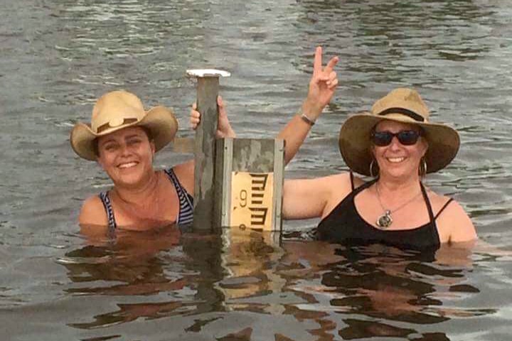 Sisters Pennie and Libby Gross stand in chest high water next to a post with a water level on it.