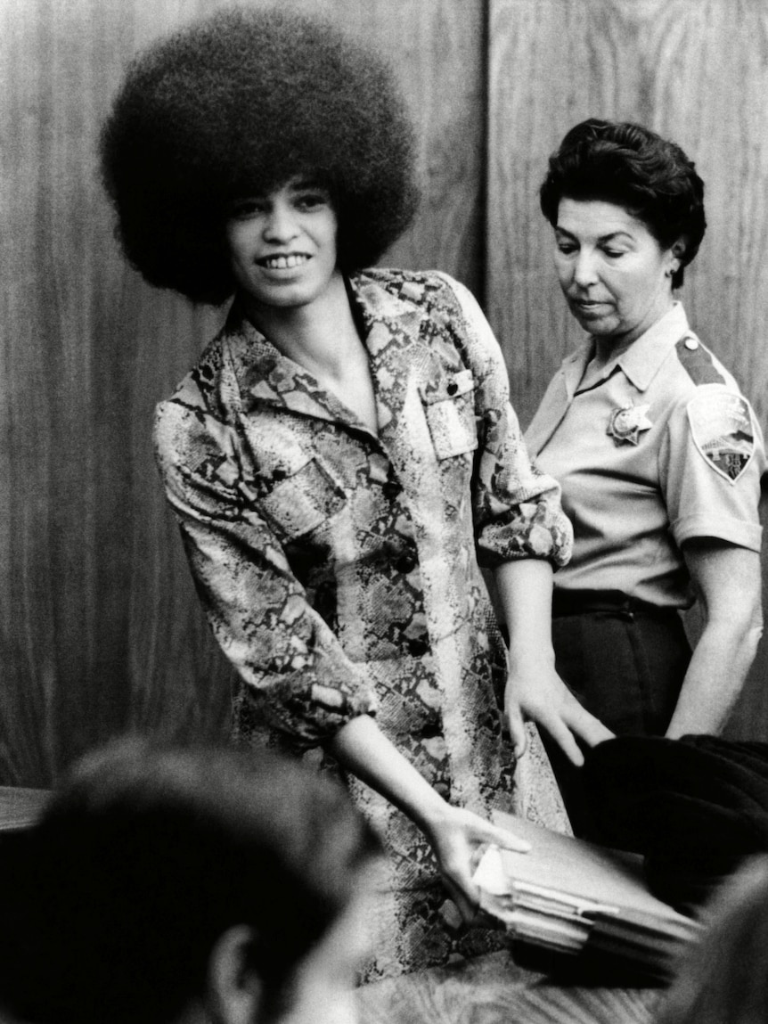 A black and white 70s photo of a Black woman with an afro, Angela Davis, she gathers documents as a white court officer looks on