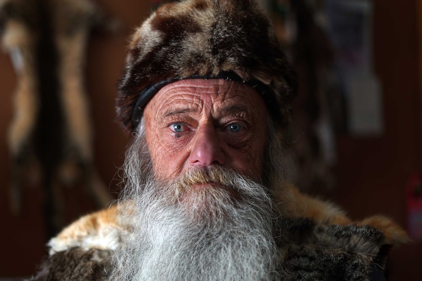 A man wearing a feral cat fur hat with a white beard stares blankly down the camera