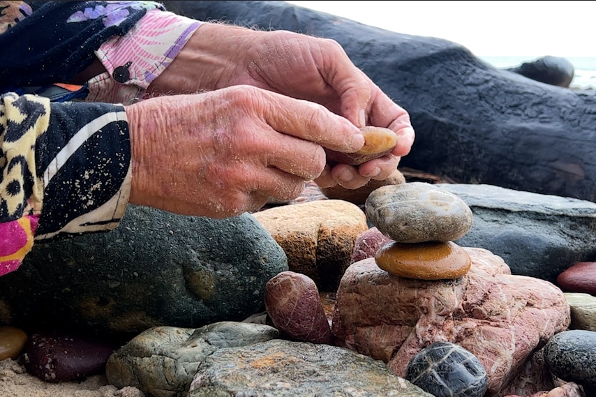 An older mans hands, placing colourful stones in a pile on a beach.