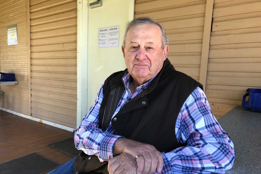 An older man wearing white, blue and pink check shirt, black sleeveless jacket sits at table in front of shuttered building. 