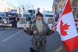 A bearded man stands in front of several semi-trailers holding Canada's flag