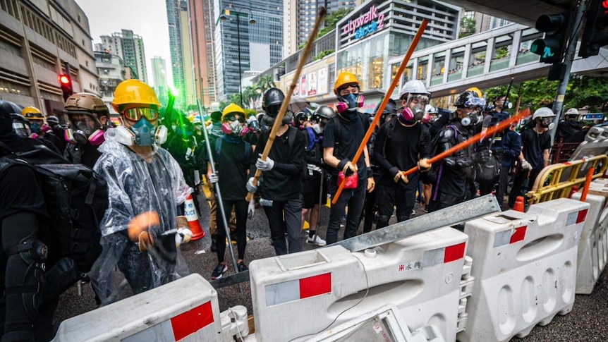 A group of people wearing black, gas masks, helmets and googles holding long poles stand behind barricades.
