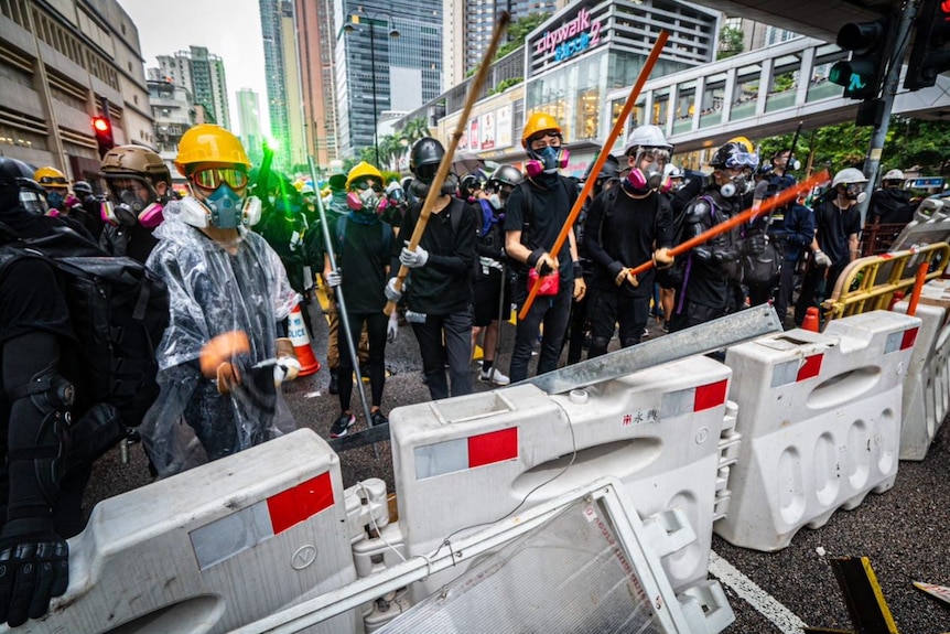 A group of people wearing black, gas masks, helmets and googles holding long poles stand behind barricades.