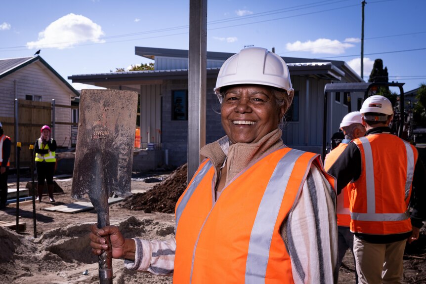 A smiling Indigenous woman in a hard hat and high-vis stands in front of a construction site, holding a shovel.
