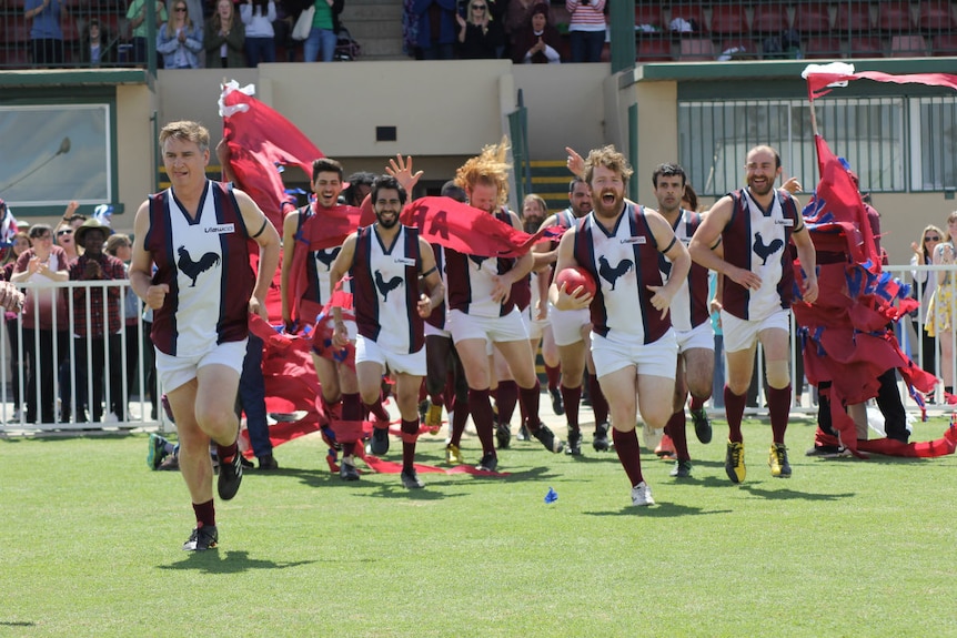 A group of men in AFL gear run through a banner and onto the footy field.