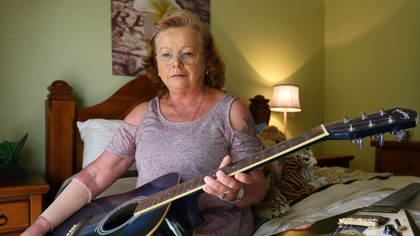A woman sits on a bed holding an acoustic guitar. An electric guitar sits on the bed beside her.