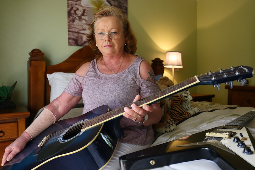 A woman sits on a bed holding an acoustic guitar. An electric guitar sits on the bed beside her.