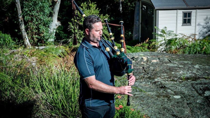Jeremy plays the bagpipes while standing in front of gum trees and a white shed.