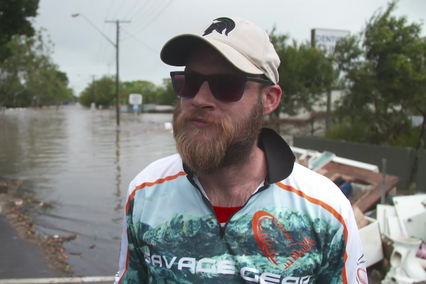 Man with a beard wearing a cap, sunglasses and activewear with floodwaters on the road behind him.