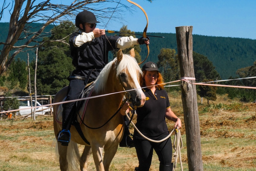 Woman on horseback aiming bow and arrow while being led by another woman.