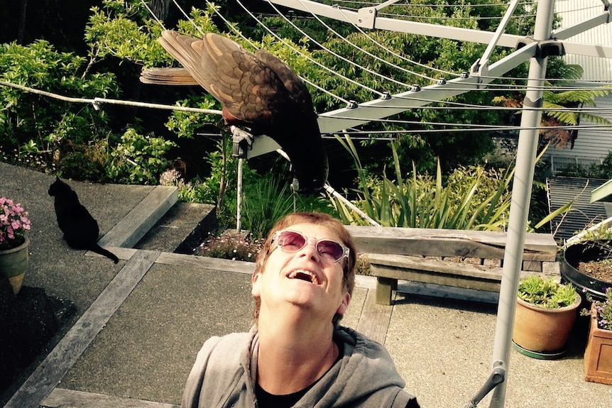 A woman wearing sunglasses looking up and smiling at a parrot that is hanging of a clothesline and leaning towards her face.