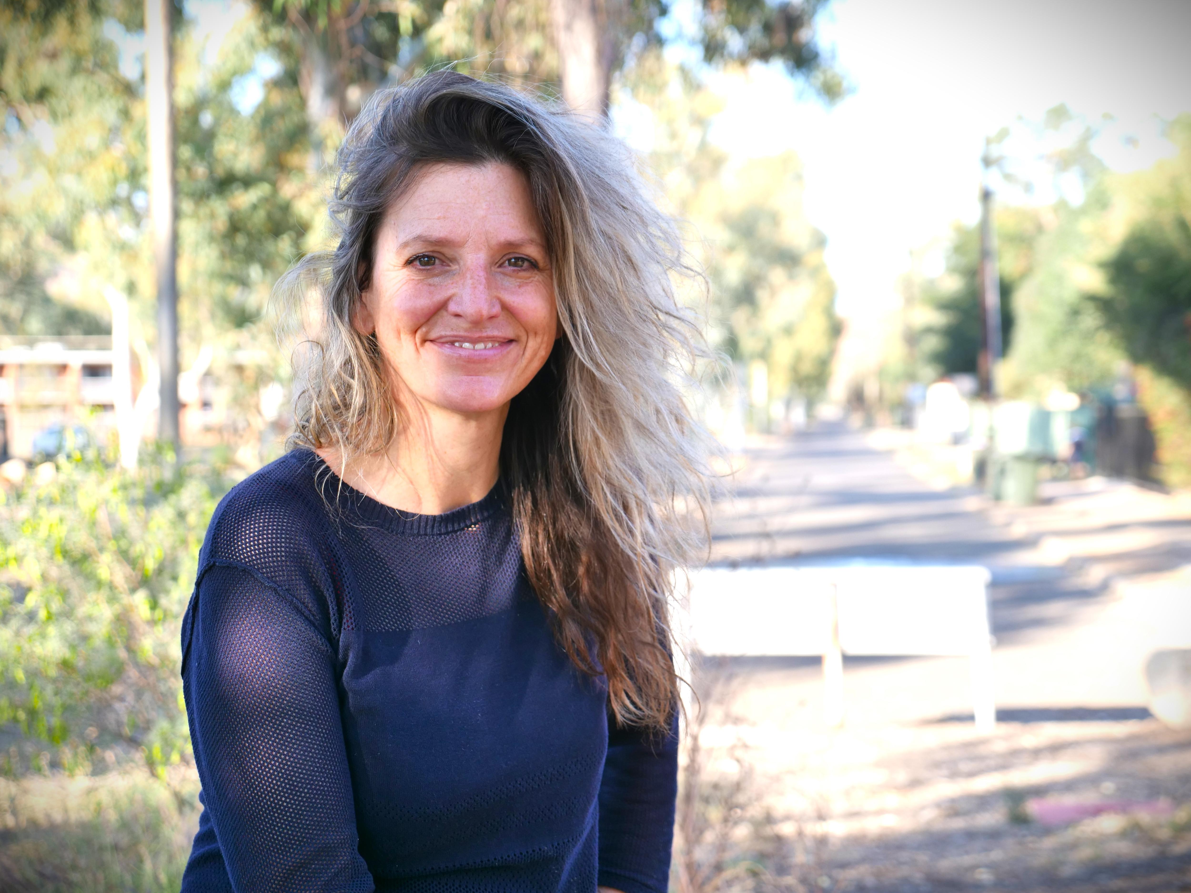 A smiling woman with bushy blonde hair, dark blue top, sits in an outdoor setting, sunny, blurred background. 