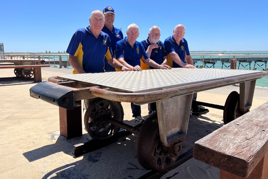 Five men wearing blue polo shirts sit at a picnic table with railway wheels on the bottom in front of a jetty and sea