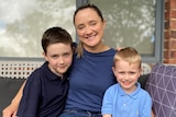 A woman sits in an outdoor lounge with her arms around two young boys, they are all smiling