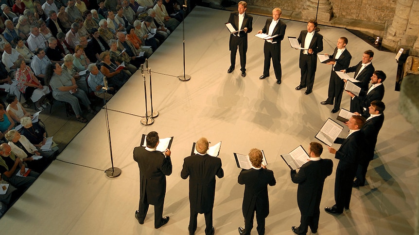 A group of male singers in tuxedos perform on stage in a semi-circle. Viewed from above with the front rows of audience watching