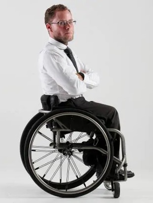 A man in a wheelchair wearing a black and white suit looks at the camera.