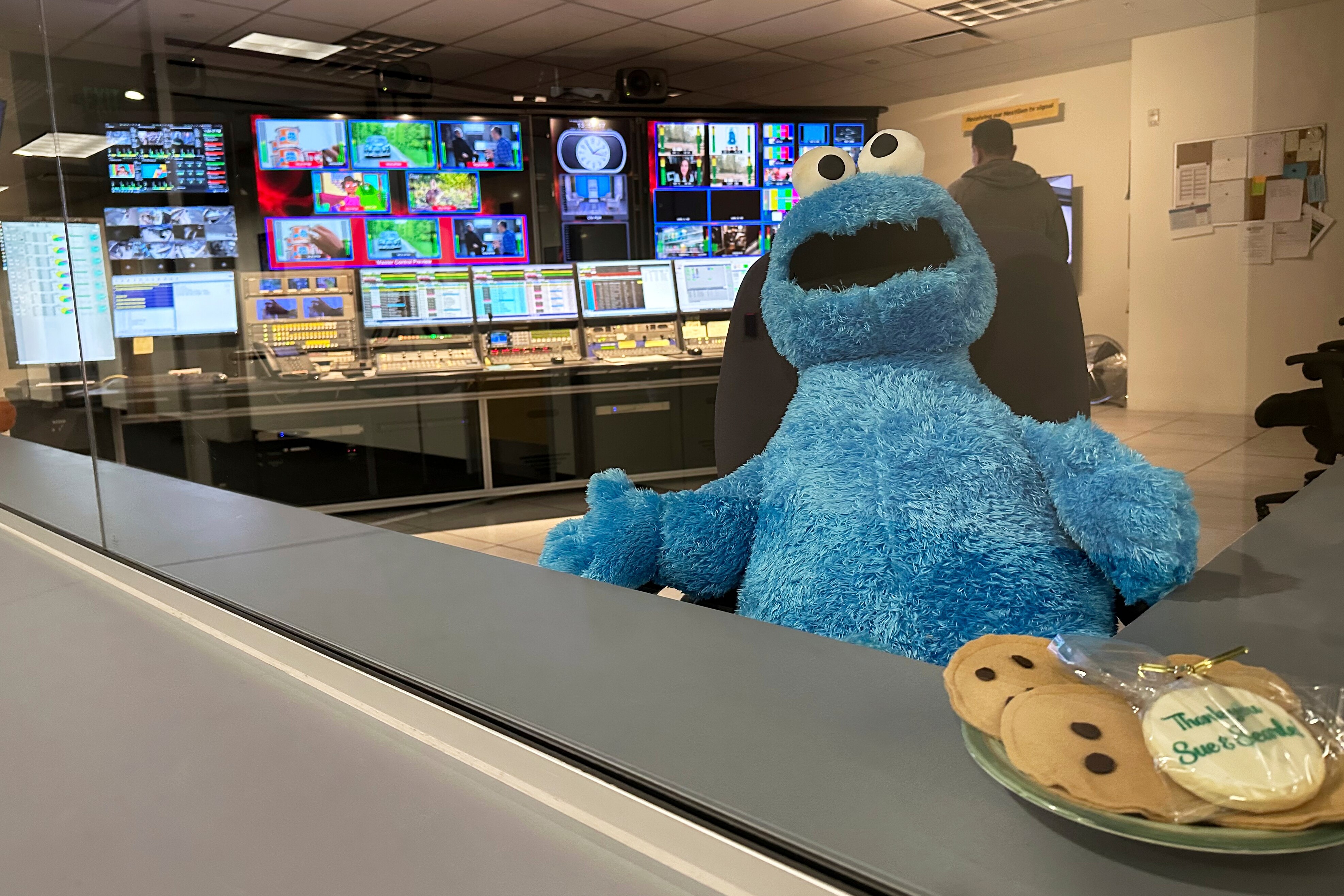 A stuffed Cookie Monster plush is seated in a television station control room in front of a plate of cookies