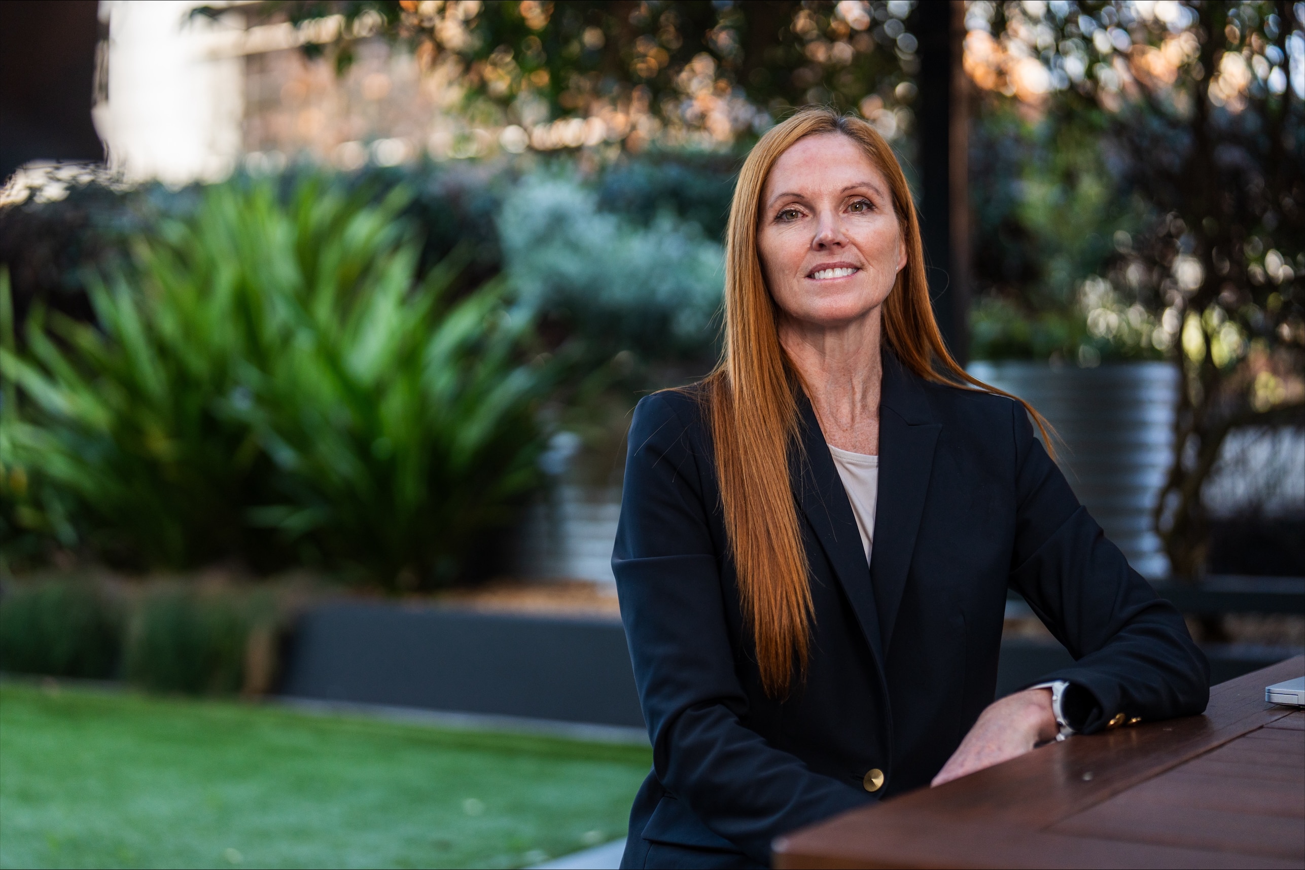 A woman leaning on a table look at the camera with a garden in the background. 