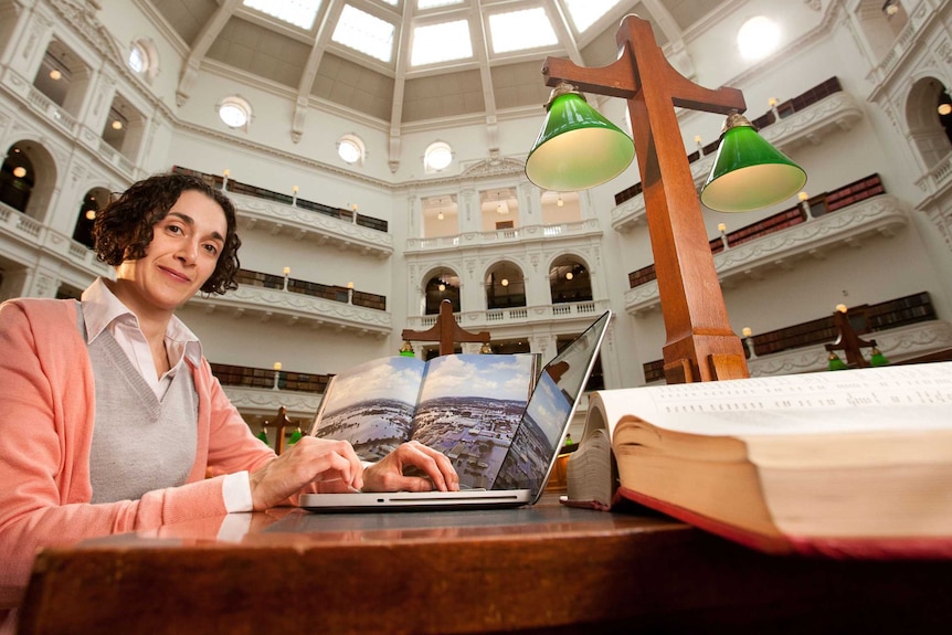 Dr Joelle Gergis sitting at a desk with a laptop and a large book in a room with a very high, vaulted roof
