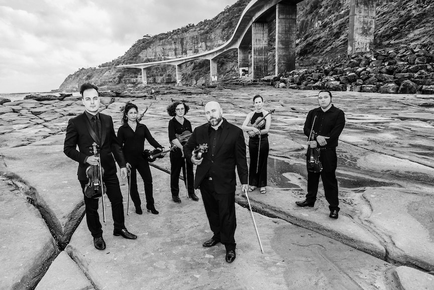 Musicians stand on an ocean rock platform wearing black and holding instruments.